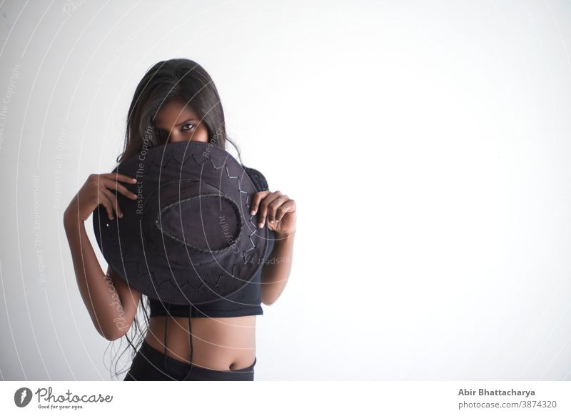 Portrait of an attractive young brunette dark skinned Indian woman in western black dress holding a cowboy hat in front of her face standing in white studio background.