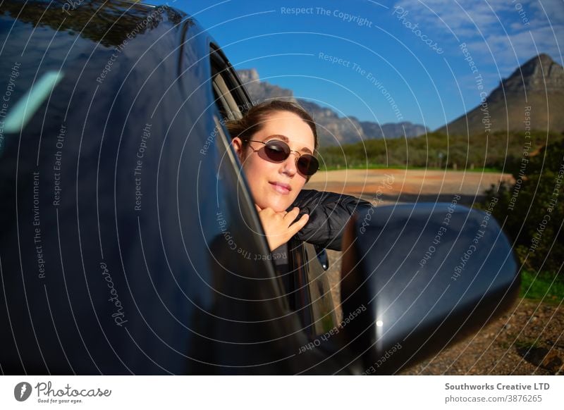 Young Woman On Road Trip Vacation Leaning Out Of Rental Car Window With Mountain Landscape Behind car rental road trip woman car hire young women holiday