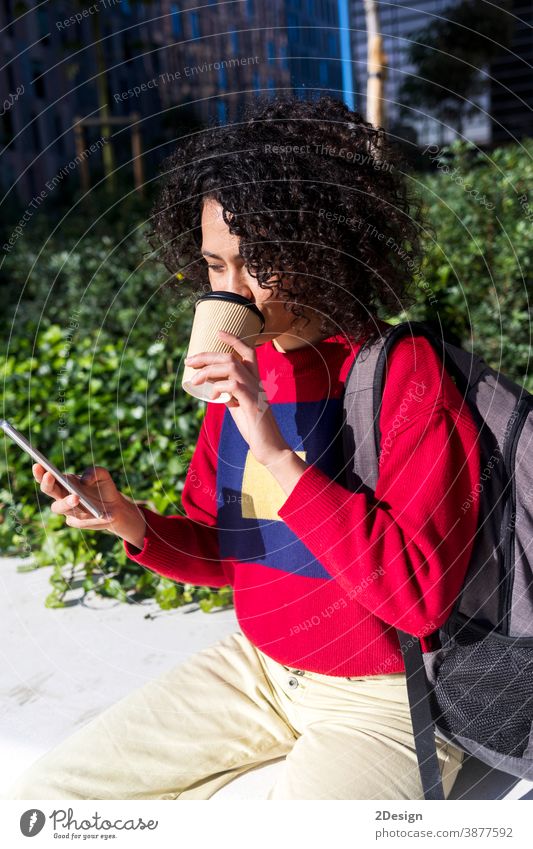 Beautiful young afro woman sitting on a bench outdoors, using mobile phone while drinking takeaway coffee cup smartphone take-away girl female backpack outside