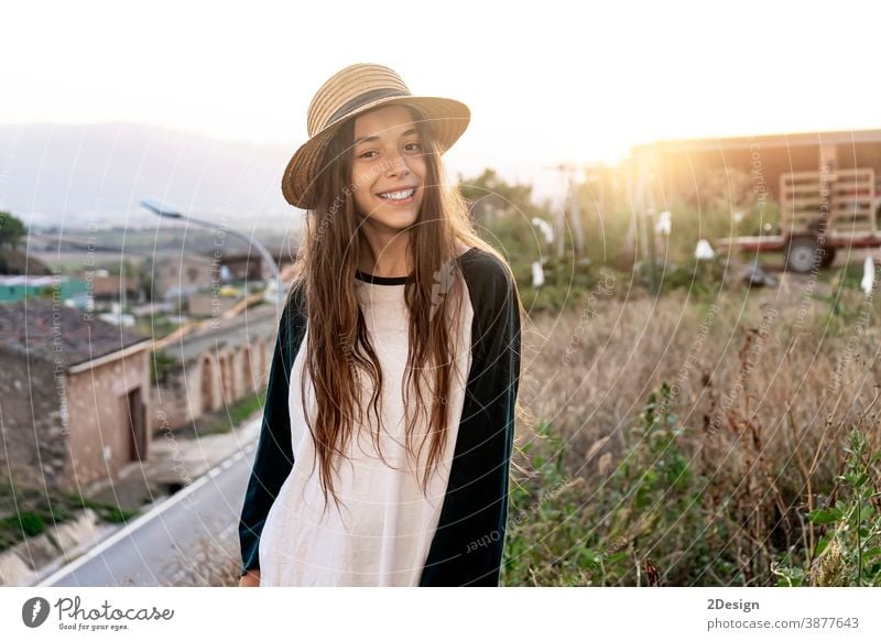 Young smiling teenager traveler girl with summer clothes and hat standing outdoors while looking at camera happy toothy long hair outside young youth laughing