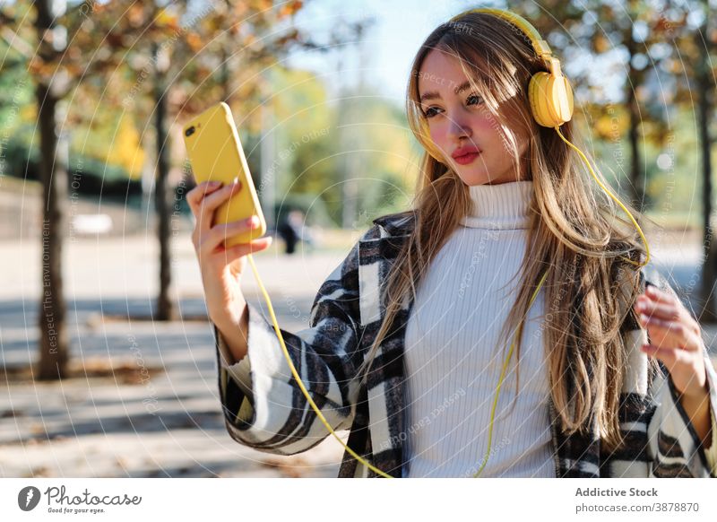Trendy young woman with yellow headphones and smartphone in autumn park selfie using colorful trendy style millennial female checkered music listen wireless