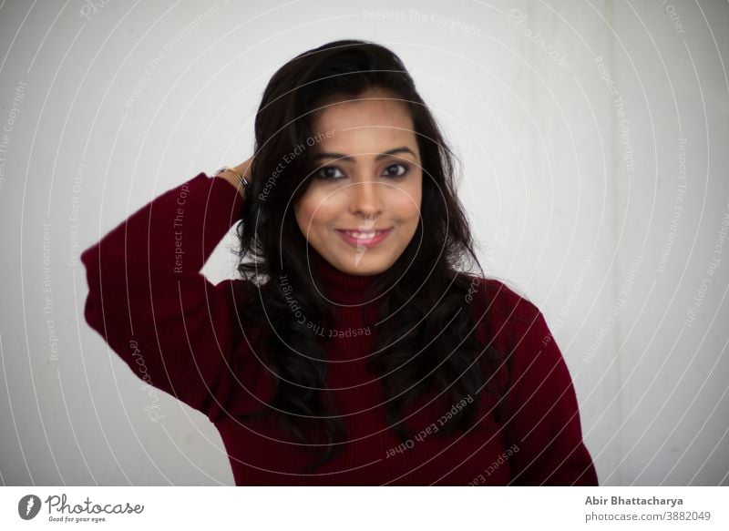 Portrait of young smiling brunette Indian Bengali woman in maroon western woolen high neck sweater leaning to wall in white copy space background. Indian lifestyle and winter collection