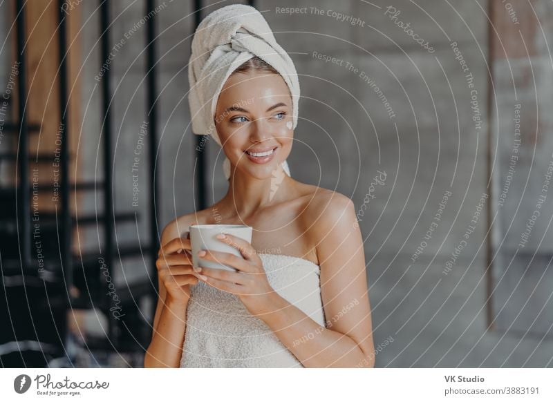 Attractive young woman puts moisturizing cream on face after showering, smiles broadly, wrapped in towels, drinks coffee, poses indoor, thinks about something pleasant. Beauty skin care concept