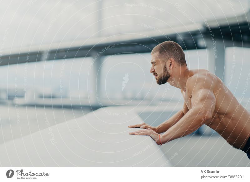 Sideways shot of handsome muscular man with thick beard does push ups exercise has workout outdoor leans on bridge fence engaged in physical activity poses with bare torso has strong muscles