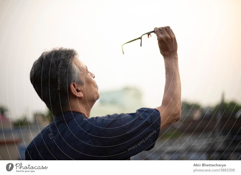 An old / aged Indian Bengali man in blue shirt is holding his spectacles up on the air to watch it carefully while standing on a rooftop under the open sky. Indian lifestyle and seniors