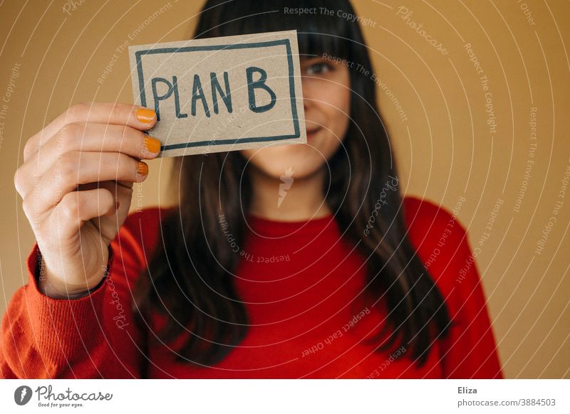A dark-haired woman in a red sweater holds up a sign saying Plan B Woman Flexibility alternative Planning Business Human being second way of education