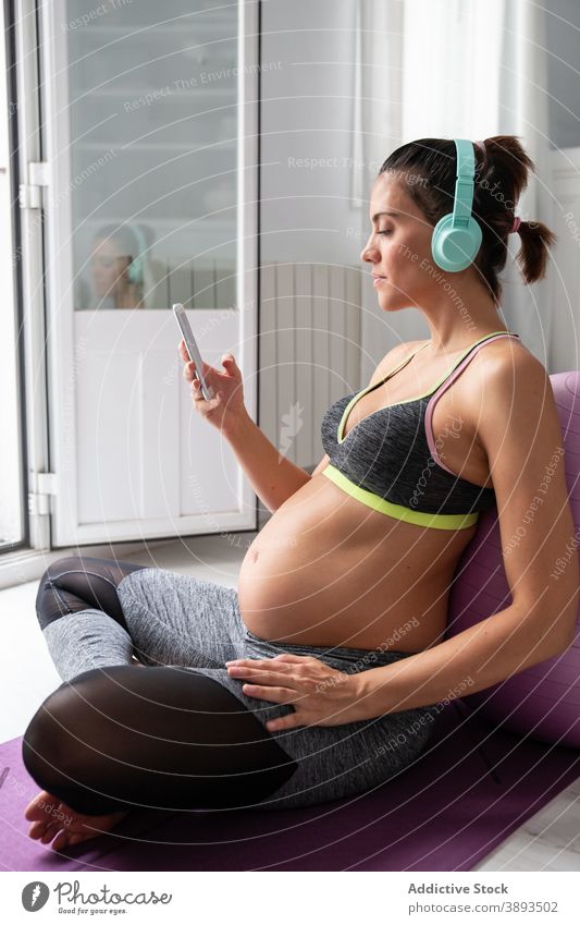 Pregnant woman in sportswear on mobile phone at home pregnant yoga pregnancy smartphone mat belly listen music female using headphones gadget wellness relax