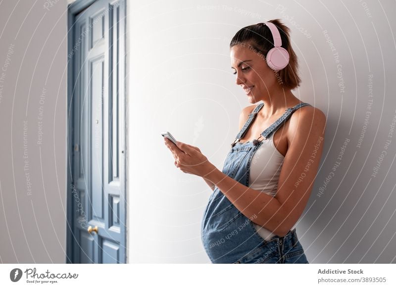 Pregnant woman enjoying music in headphones at home pregnant listen pregnancy overall song wireless female smile cheerful room melody sound playlist happy tune