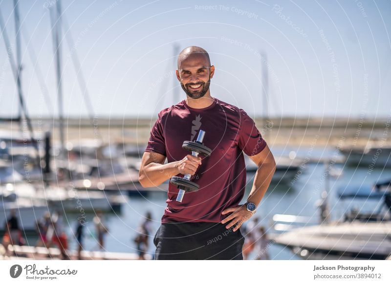 A man standing with weights outdoor by the seaside portrait exercise sport park urban day sunny young copy space marina boat serious trainer training personal