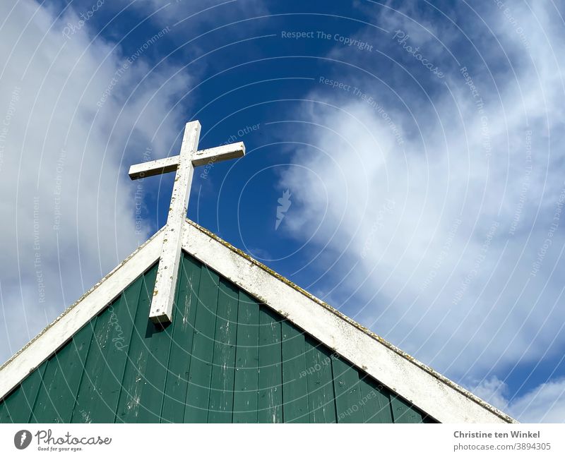 White wooden cross on the gable wall of a chapel, which is covered with green and white wood.  Everything is somewhat weathered. View from below against the blue cloudy sky. Spiekeroog I colour combination