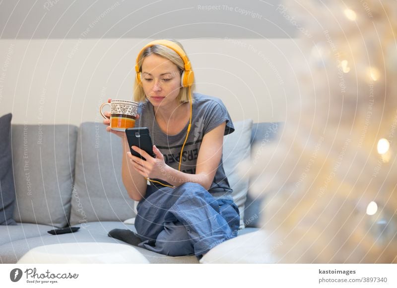 Young cheerful woman sitting indoors at home living room sofa using social media on phone for video chatting and staying connected with her loved ones. Stay at home, social distancing lifestyle