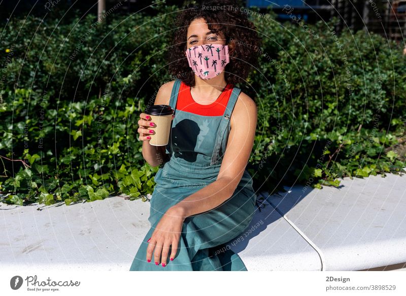Protected woman with curly hair is sitting in a sunny day while looking at camera and wearing a special mask virus disease epidemic protection pandemic