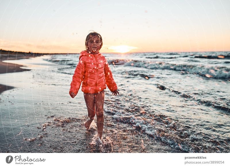 Playful little girl splashing a water towards camera enjoying a free time over sea on a sand beach at sunset excited positive emotion carefree nature outdoors