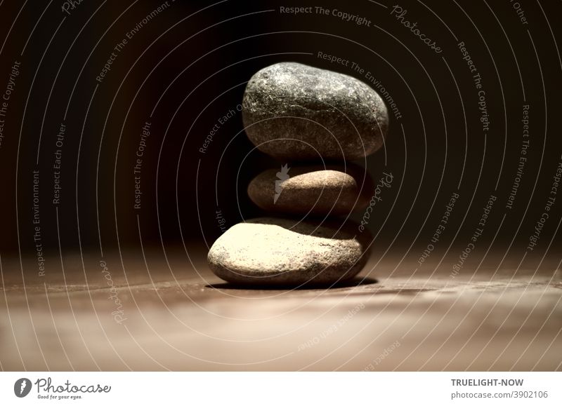 Three pebbles lie in balance on a dimly lit table in front of a dark background, as if floating on top of each other, and symbolize the connection between heaven and earth or spirit and matter.