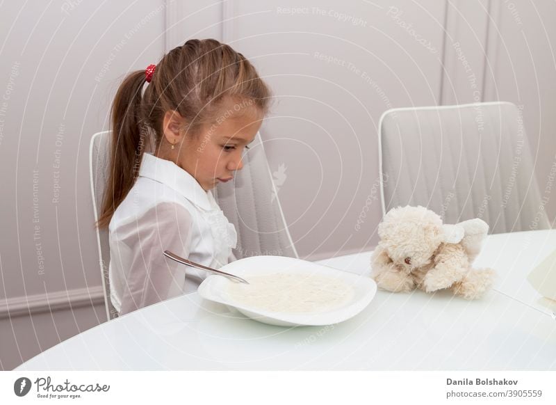 child is sitting at glass table in huge bright dining room near panoramic window. girl turned away from porridge with disgruntled face before going to school