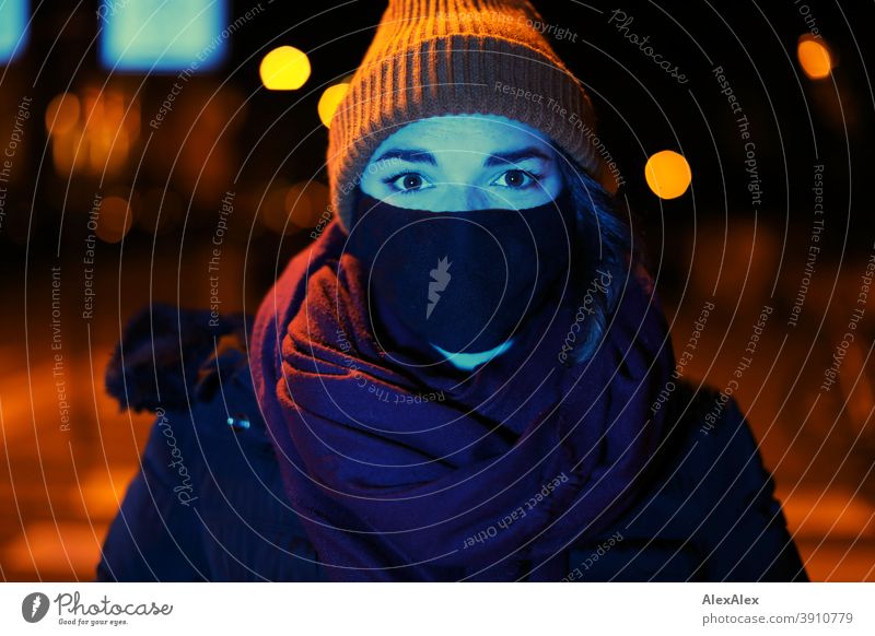 Junge Frau mit Maske und Mütze steht nachts mit blauem Licht angeleuchtet in orangem Laternenlicht in der Stadt jung Jugendliche blaues Licht Straße kariert