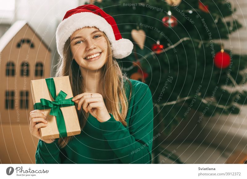 A beautiful little girl looks at the camera and smiles, holding a kraft paper gift tied with a green ribbon in her hands. Christmas mood. Concept for New Years holiday at home.