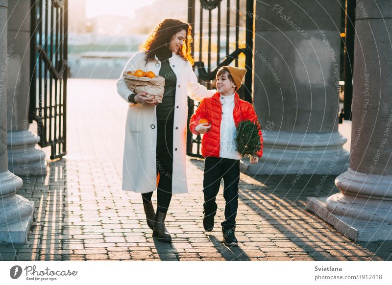 A beautiful mother with her son in a bright orange jacket and a fashionable yellow hat are walking from the store holding a package of oranges and a small Christmas tree. Shopping for the holiday. Preparing for Christmas and New Year