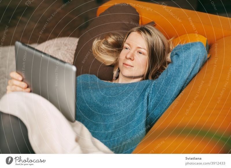 Happy girl lying on a comfortable sofa in the living room, watching a movie on a tablet. A young woman has a rest and communicates with friends using a tablet. Work online, freelance, social distance