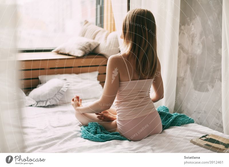 A young woman in pyjamas is sitting on a bed in a Lotus position with her fingers folded in a mudra gesture, enjoying a morning of deep mindfulness meditation. Rear view