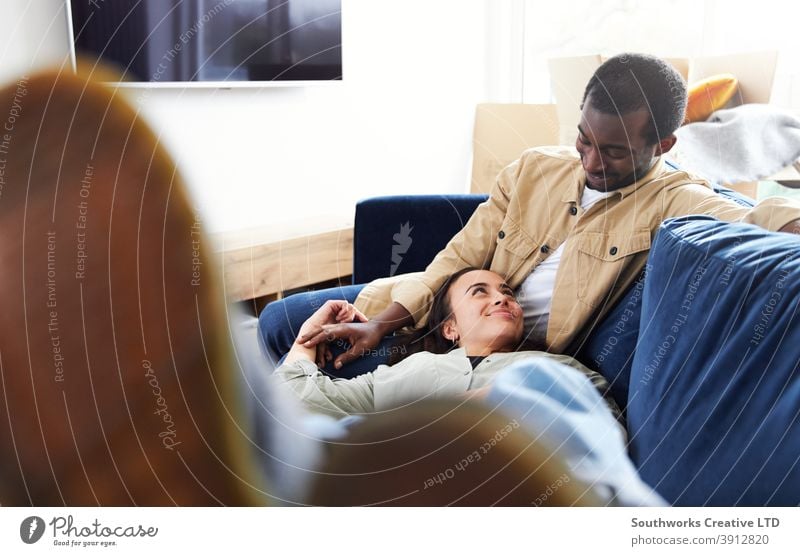 Young Couple In New Home Lying On Sofa In Lounge On Moving Day Surrounded by Removal Boxes couple young couple house buying sitting lying sofa living room