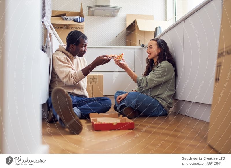 Young Couple Celebrate Moving Into New Home Sitting On Floor Eating Pizza In Kitchen With Removal Boxes couple young couple house buying pizza takeaway