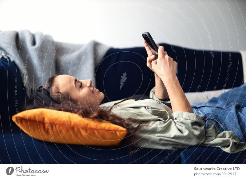 Young Smiling Woman Relaxing At Home Lying On Sofa Checking Social Media On Mobile Phone woman at home lying sofa lounge mobile mobile phone cell cell phone
