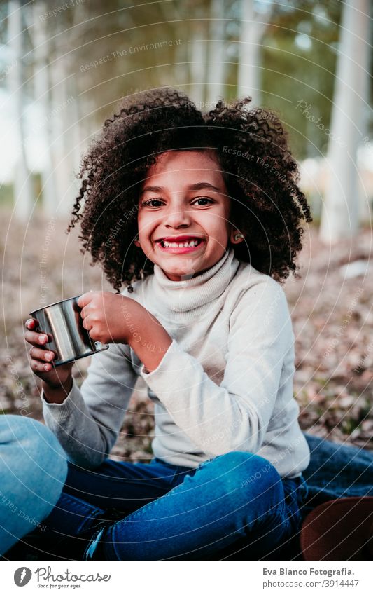 portrait of cute afro kid girl outdoors doing picnic, holding mug of water, autumn season, beautiful trees background nature golden hour hat brown leaves