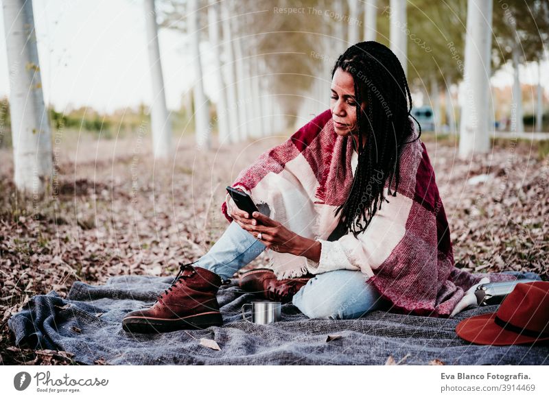 portrait of hispanic mid adult woman outdoors holding wrapped in blanket. Using mobile phone during picnic.Autumn season autumn afro woman latin sunset nature