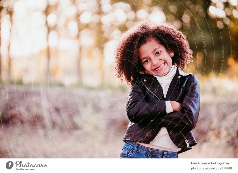 portrait of cute afro kid girl smiling at sunset during golden hour, autumn season, beautiful trees background nature outdoors hat brown leaves casual clothing