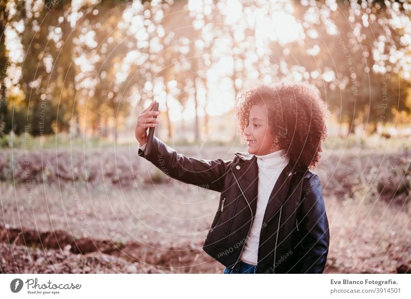 portrait of cute afro kid girl using mobile phone at sunset during golden hour, autumn season, beautiful trees background technology nature outdoors hat brown