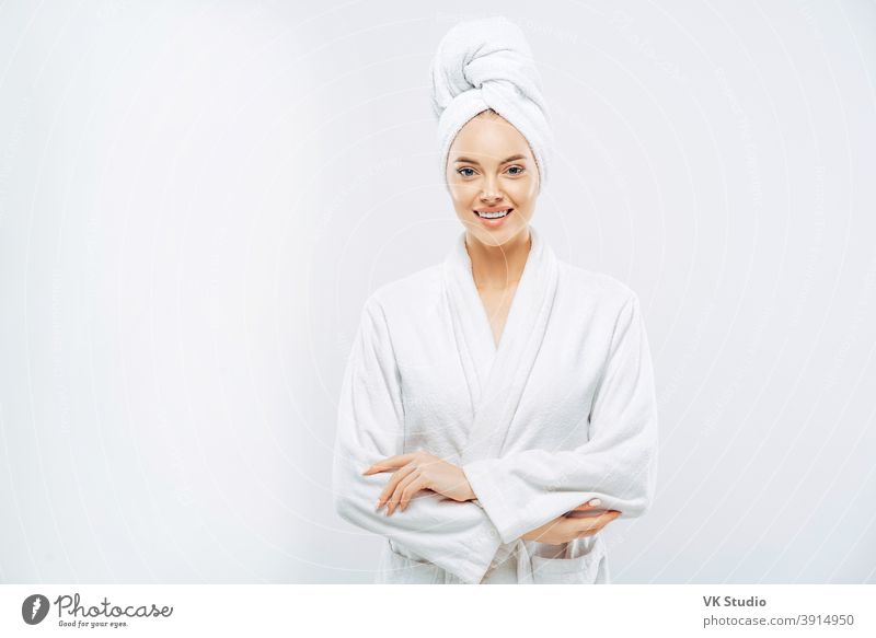 Beautiful smiling young woman has soft healthy skin after taking shower, wears bath robe and towel wrapped on head, enjoys spare time at home, isolated over white background. Wellness concept.