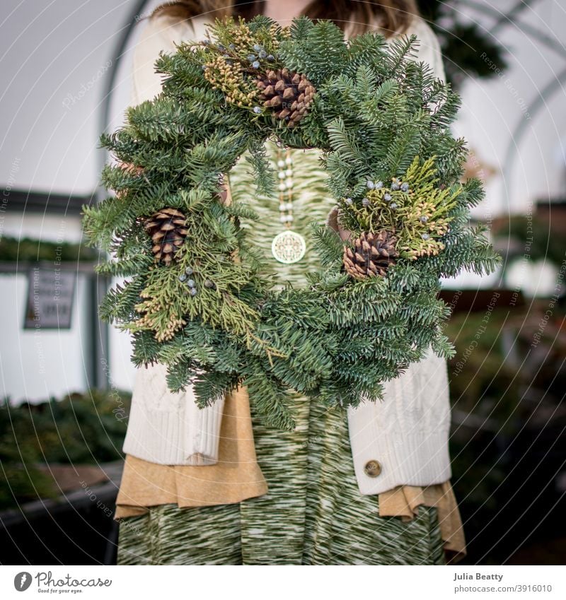 Woman holding Evergreen wreath with pine cones in front of body; inside of greenhouse christmas circle swag tree farm decoration decorate garland holiday x-mas