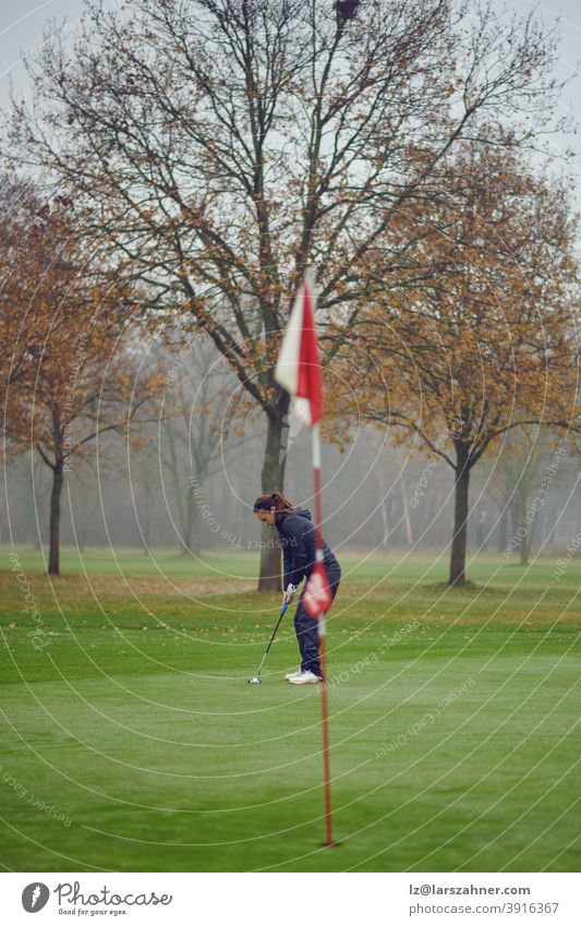 Middle-aged fit woman golfer putting for the hole on the green viewed from the flag concentrating for the next swing winter autumn windy misty middle-aged