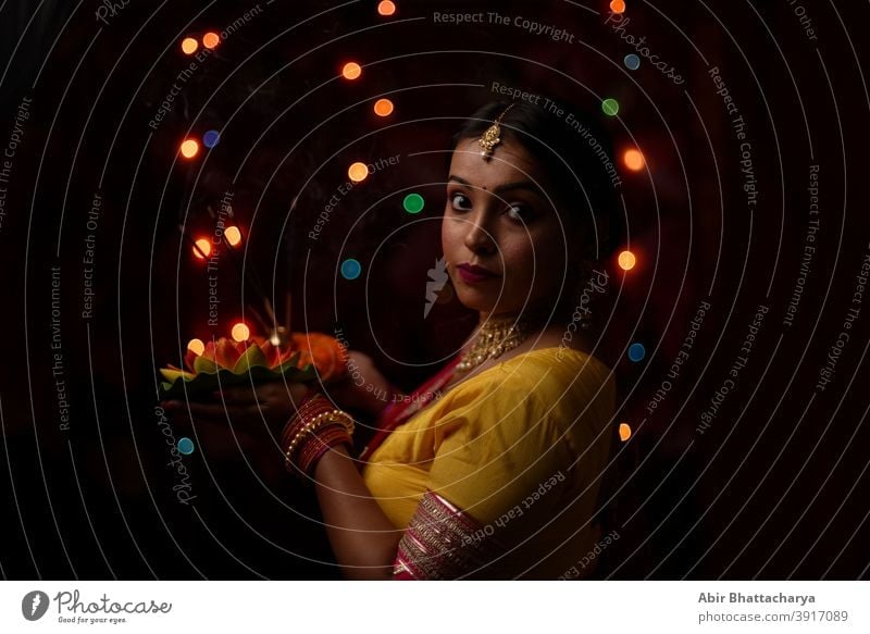 An young and beautiful Indian Bengali woman in Indian traditional dress is holding a Diwali diya/lamp in her hand in front of colorful bokeh lights. Indian lifestyle and Diwali celebration