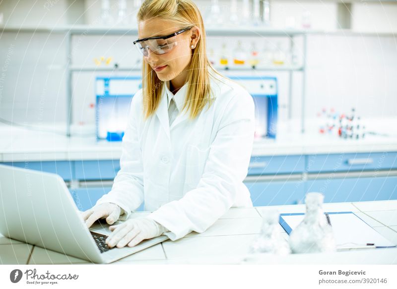Female researcher in white lab coat and protective goggles using laptop while working in the laboratory analyzing background bio chemical chemist clinic