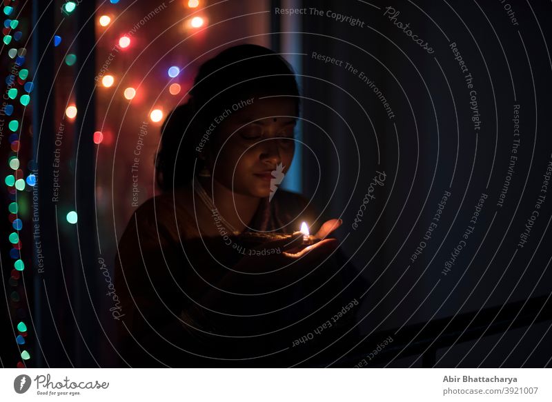 An young and beautiful Indian Bengali woman in Indian traditional dress is holding a Diwali diya/lamp in her hand standing on a balcony in darkness. Indian lifestyle and Diwali celebration