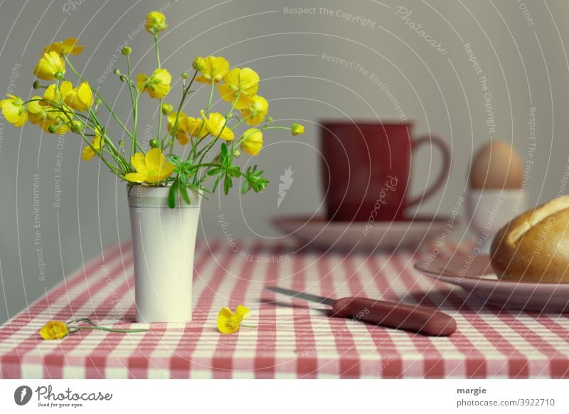 A vase with buttercups stands on a breakfast table with a red checked tablecloth. A roll on a plate with a knife, in the background a red cup on a saucer and a boiled egg in an egg cup