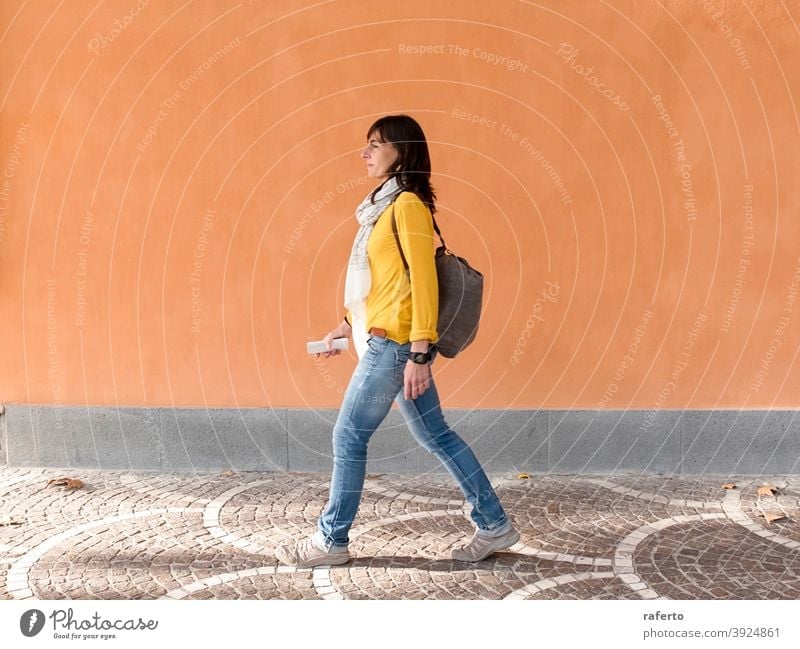 Side view of a backpacker traveler walking against orange wall in the city woman street outdoor person urban 1 holiday maker brunette lady vacation trip tourism