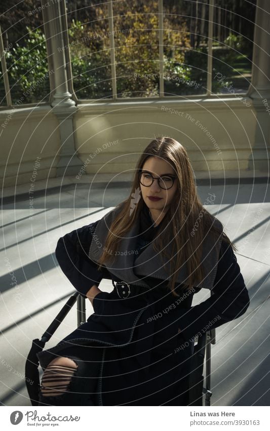 A gorgeous girl dressed in a fashionable autumn coat is sitting on a chair in the middle of the Crystal Palace. In other words, Palacio de Cristal is based in Retiro Park, Madrid. Pretty face brunette model is also wearing glasses.