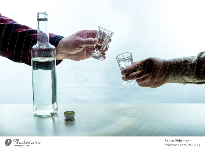 On a table there is a bottle with alcohol. The hands of two men are holding shot glasses. They toast with each other. Alcoholic drinks alcoholism Addiction
