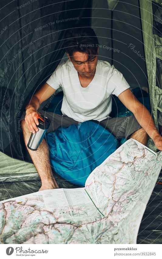 Young man looking at a map of mountain trails planning next trip on summer vacation sitting in a tent on camping examining tourist travel copy space wanderlust
