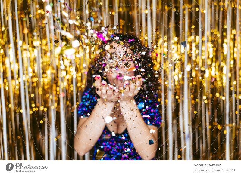 Selective focus. A woman in an elegant shiny dress and Christmas decorations behind her blowing some confetti to the camera. New Year's Eve party concept