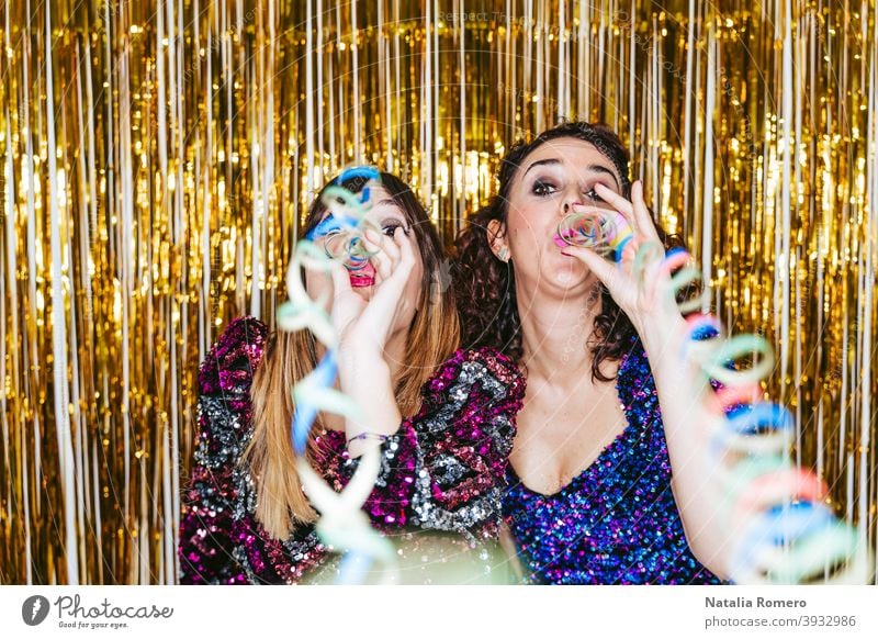 Two beautiful women in elegant clothes with Christmas decorations behind them blowing streamers to the camera while celebrating in a New Year's party. New Year's Eve party concept