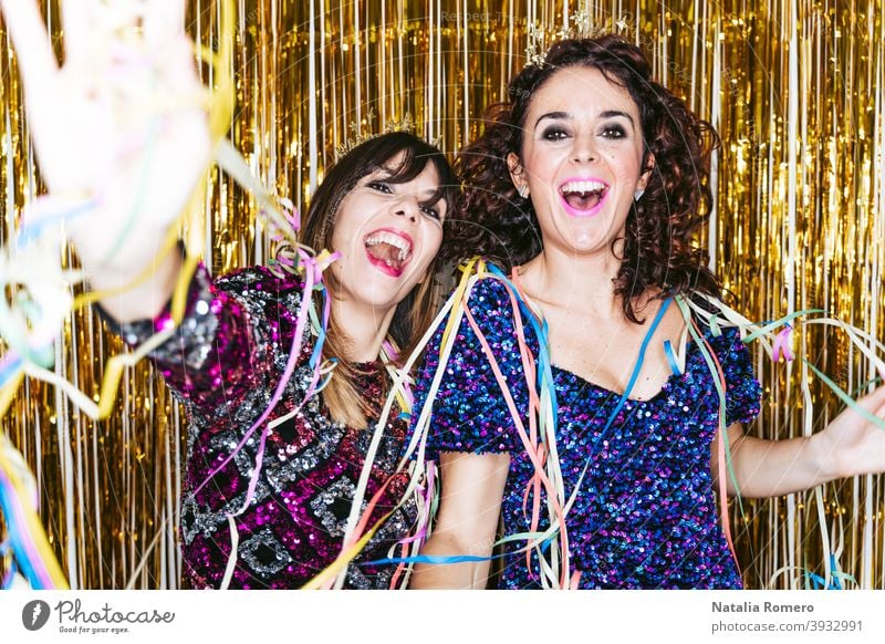 Two beautiful brunette women in elegant clothes and decorations behind them celebrating the end of the year together while playing with some streamers. New Year's Eve party at home concept