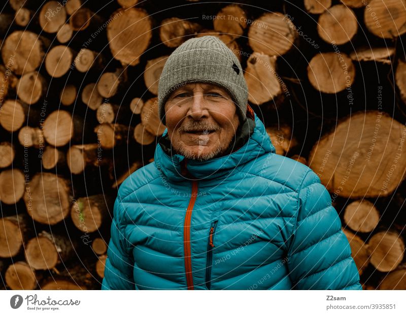 Portrait eines sportlichen Rentners vor einem Holzstapel spazieren natur outtdoor rentner alter mann portrait mütze winter kälte landschaft wald sträucher isar