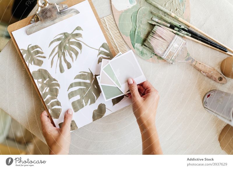 Crop artist with paper color palette and painting in workshop pastel tone monstera sample craftswoman female artisan green table studio creative hobby handmade