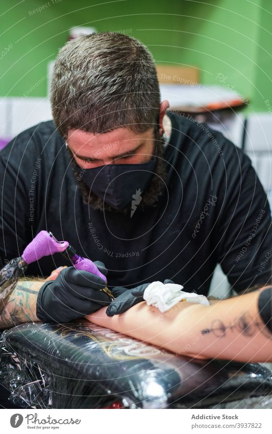 Focused master making tattoo on arm of client man tattooist work create concentrate ink instrument machine salon professional occupation male beard casual mask