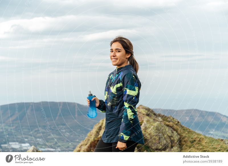 Happy young ethnic woman drinking water during training in mountainous valley sportswoman workout trekking smile thirst wellness wellbeing fitness healthy