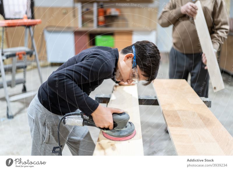 Focused child polishing wood in workshop with unrecognizable grandfather boy sander machine help grandson orbital joiner craftsman occupation wooden panel